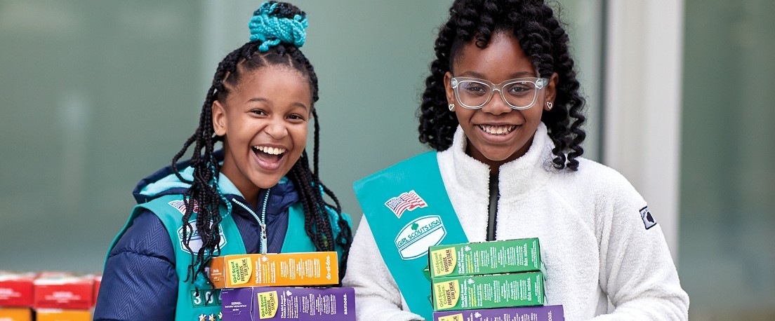 Two masked Girl Scouts outdoors holding up Girl Scout Cookie boxes