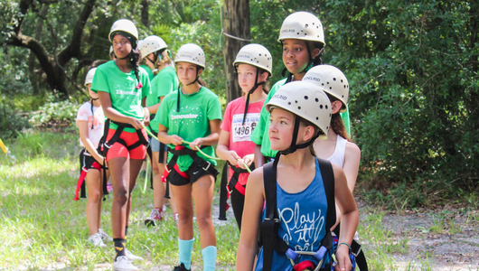 A group of Girl Scouts wearing helmets and harnesses, getting ready for the challenge course at Camp Dorothy Thomas.